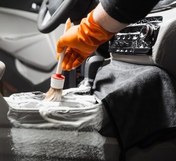 A person in an orange glove cleans a car interior with a brush and foam.